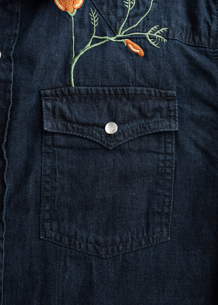 Denim shirt pocket with orange floral embroidery, featuring a silver button, showcasing intricate design on dark fabric.