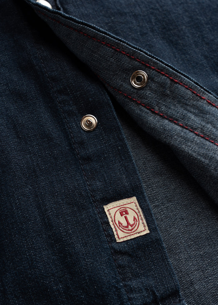 Close-up of a denim jacket with red stitching and metal snaps, featuring a small anchor patch on the inside seam.