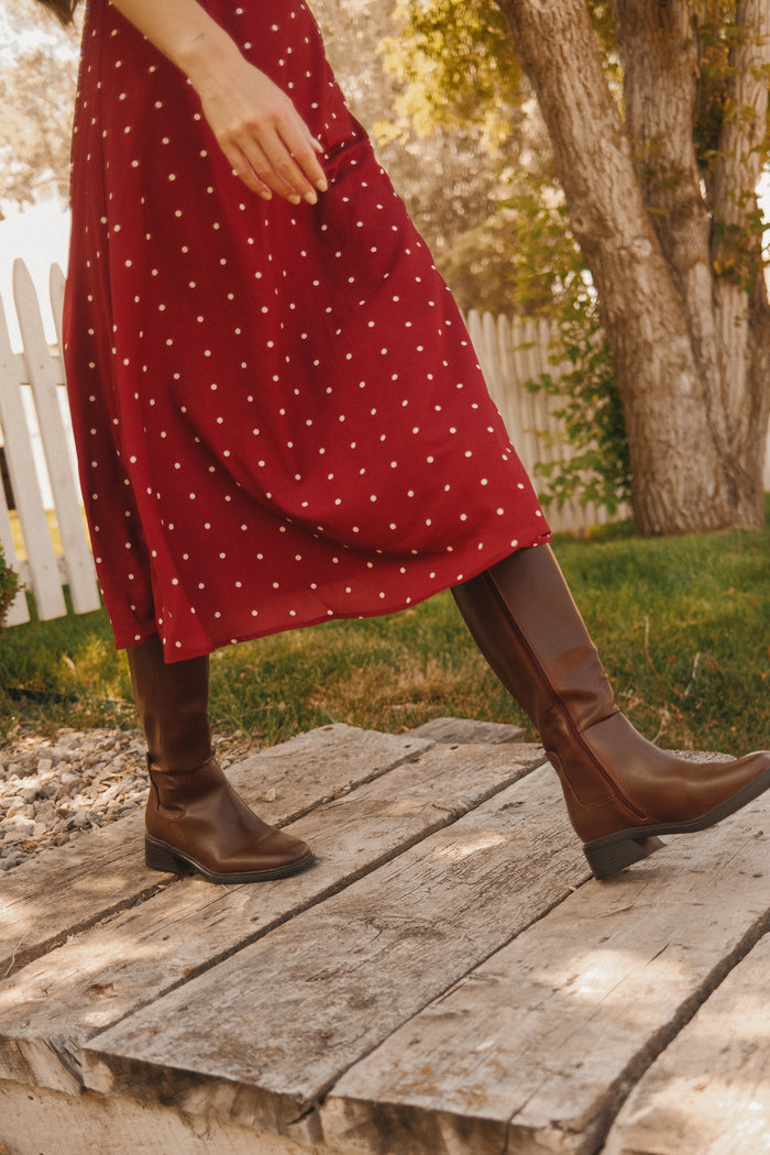 Wearing the Ivy City Co Fiona Dress in Cranberry Dainty Dot, featuring petal sleeves, and paired with brown knee-high boots, a person strolls on wooden planks outdoors amidst green grass, trees, and a white fence—a perfect addition to your fall wardrobe.