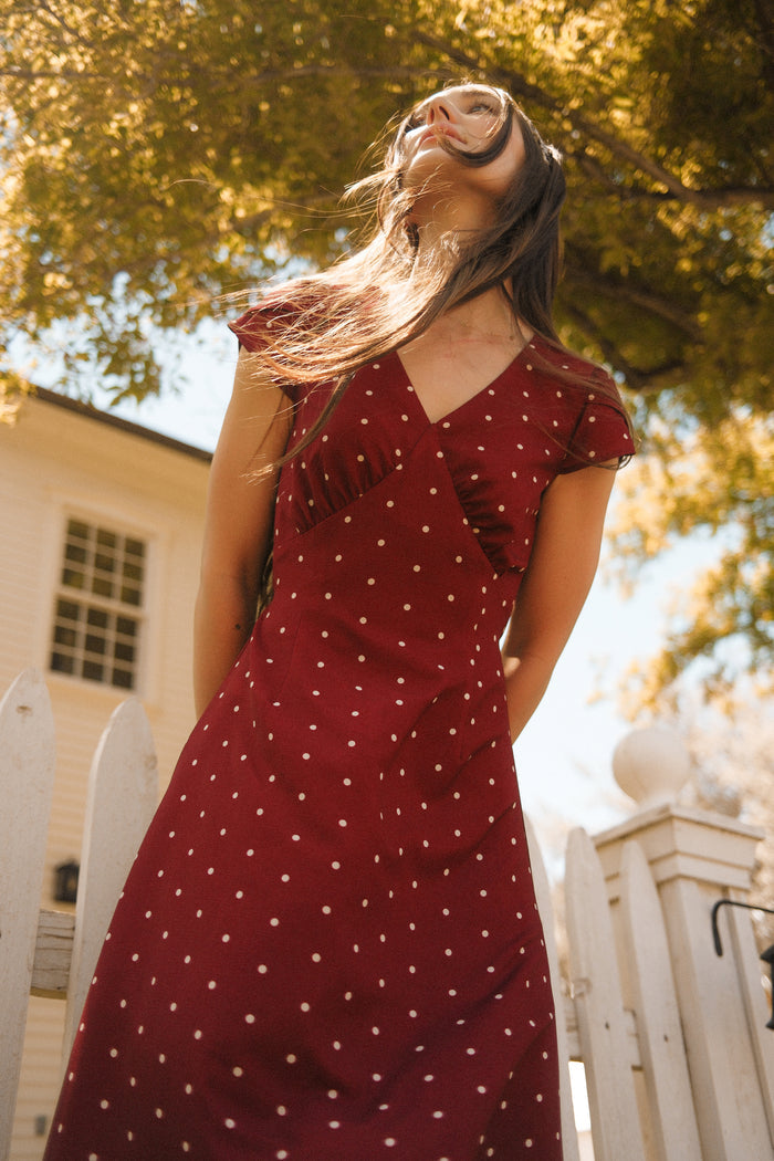 Dressed in Ivy City Co’s Fiona Dress in Cranberry Dainty Dot, featuring delicate petal sleeves, a woman stands before a white picket fence and house, gazing upward as sunlight filters through the branches—offering the perfect inspiration for your fall wardrobe.
