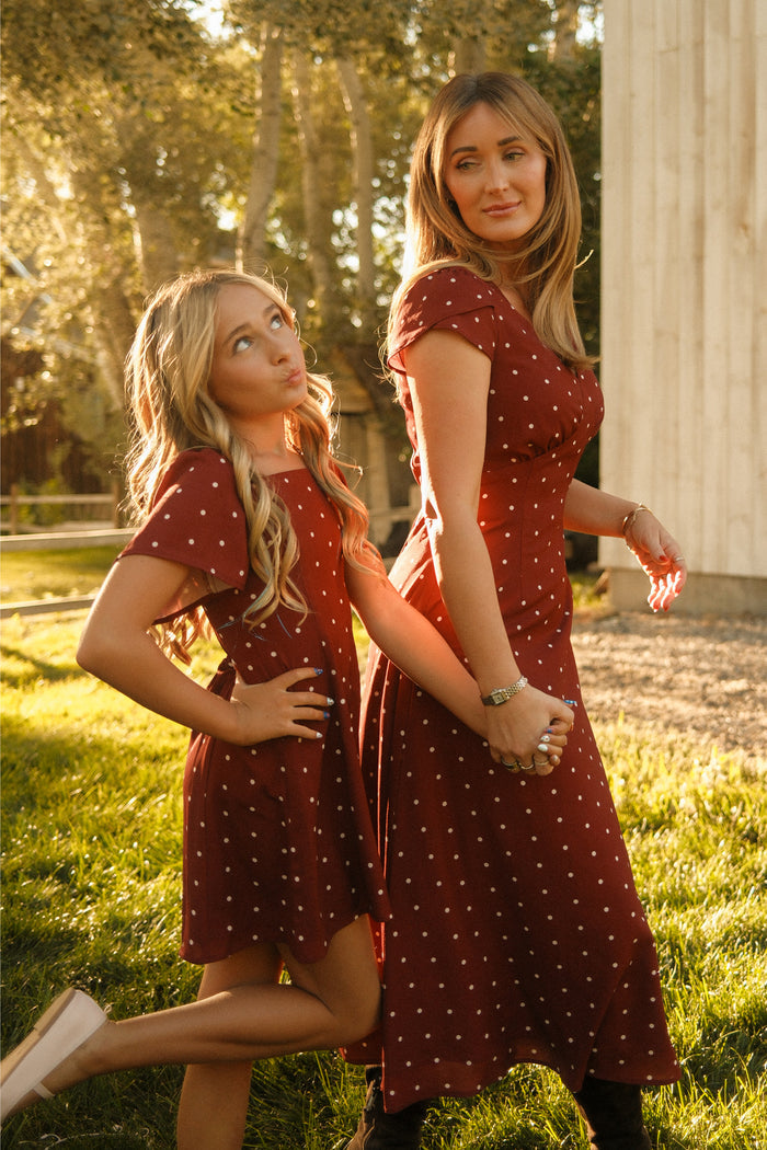 A woman and a young girl, both dressed in Ivy City Co’s Fiona Dress in Cranberry Dainty Dot featuring charming petal sleeves, hold hands and pose playfully on sunlit grass—a delightful addition to any fall wardrobe.