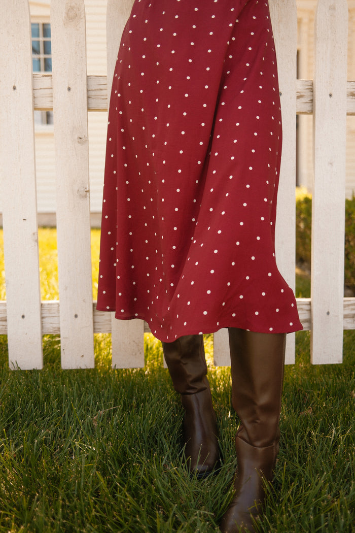 Wearing the Ivy City Co Fiona Dress in Cranberry Dainty Dot, a person stands on grass in front of a white wooden fence, their fall-ready look completed with brown boots.