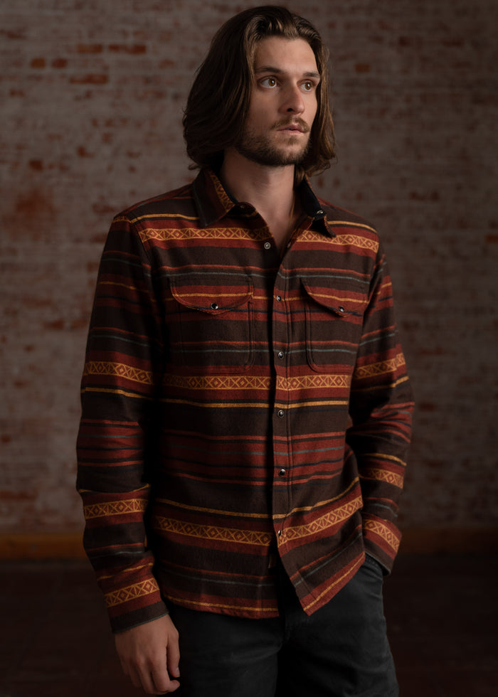 Man with long hair in a patterned brown shirt stands against a textured brick wall, looking thoughtfully to the side.