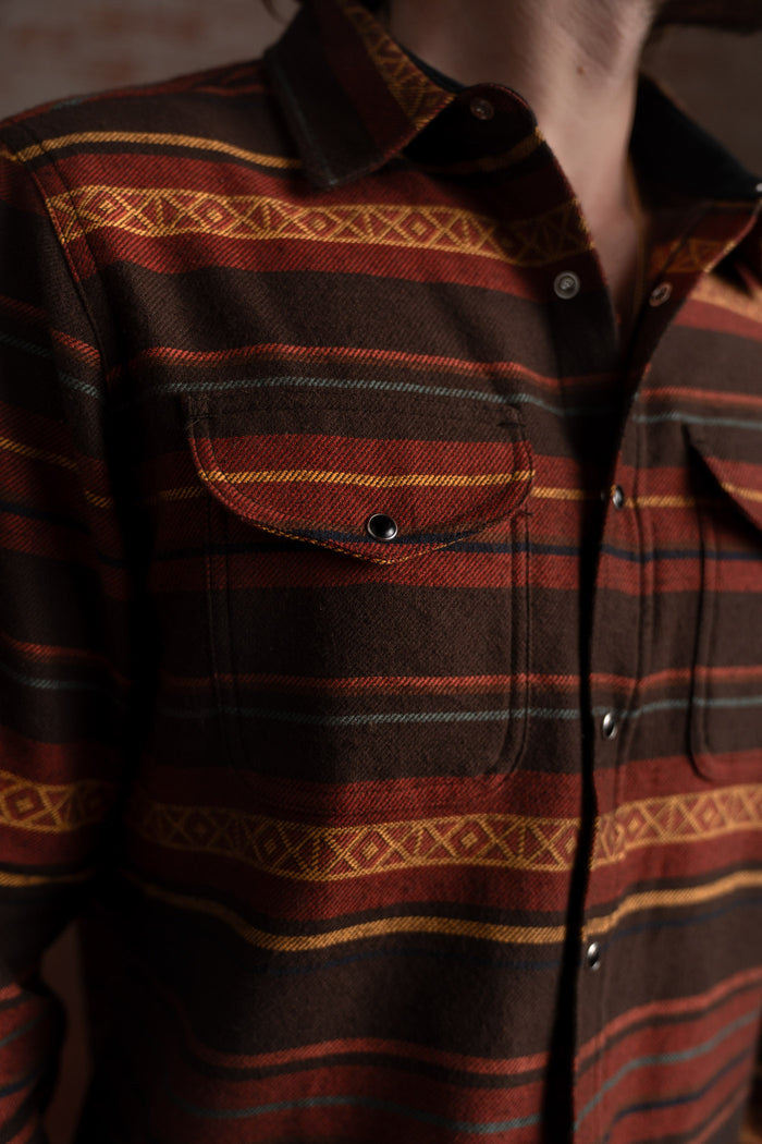 Close-up of a man wearing a brown and red striped flannel shirt with geometric patterns, highlighting the chest pocket detail.