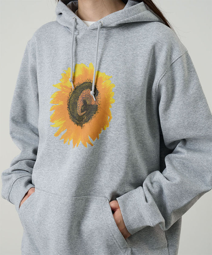 Model wearing a Gramicci heather grey G-Flower Hooded Sweatshirt with a sunflower graphic against a light-tone background.