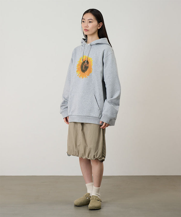 Model wearing a heather grey Gramicci G-Flower Hooded Sweatshirt with a sunflower graphic and beige skirt on a light-tone background.
