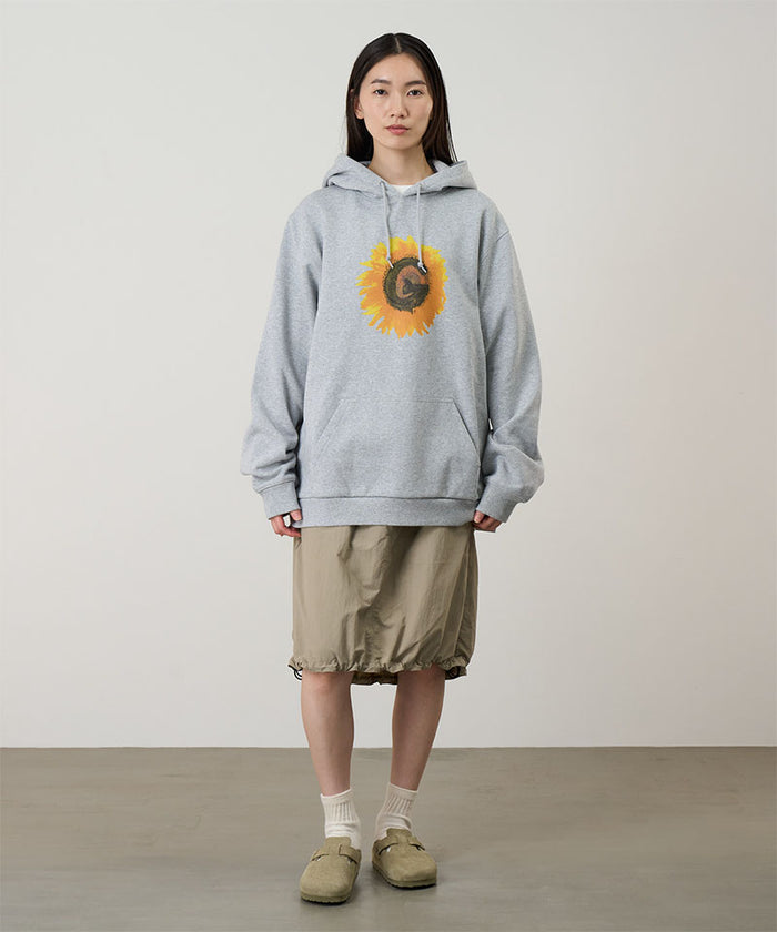 Model wearing a heather grey Gramicci G-Flower Hooded Sweatshirt with a sunflower graphic and beige skirt on a light-tone background.