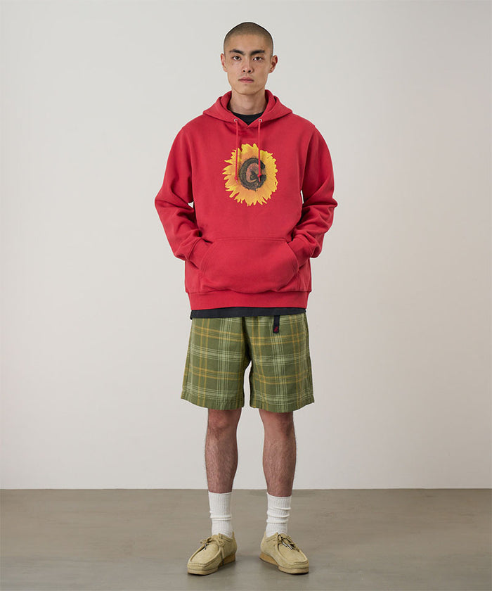 Model wearing a crimson red Gramicci G-Flower Hooded Sweatshirt with a sunflower graphic and green plaid shorts on a light-tone background.