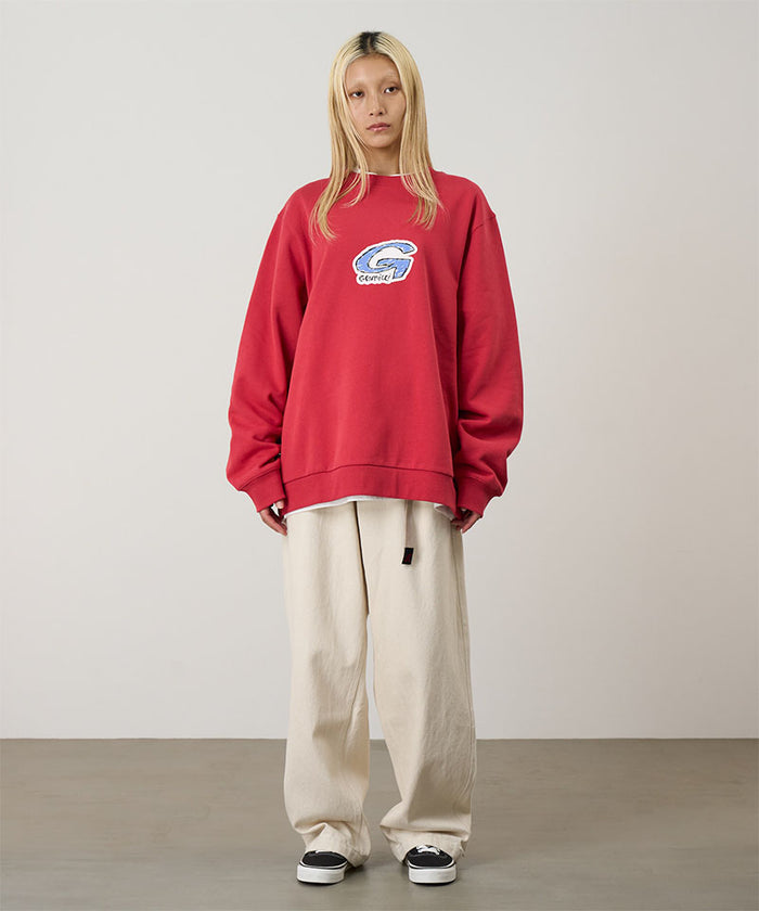 Model wearing a crimson red Gramicci G-Scribble Sweatshirt with a blue graphic and beige pants on a light tone background.