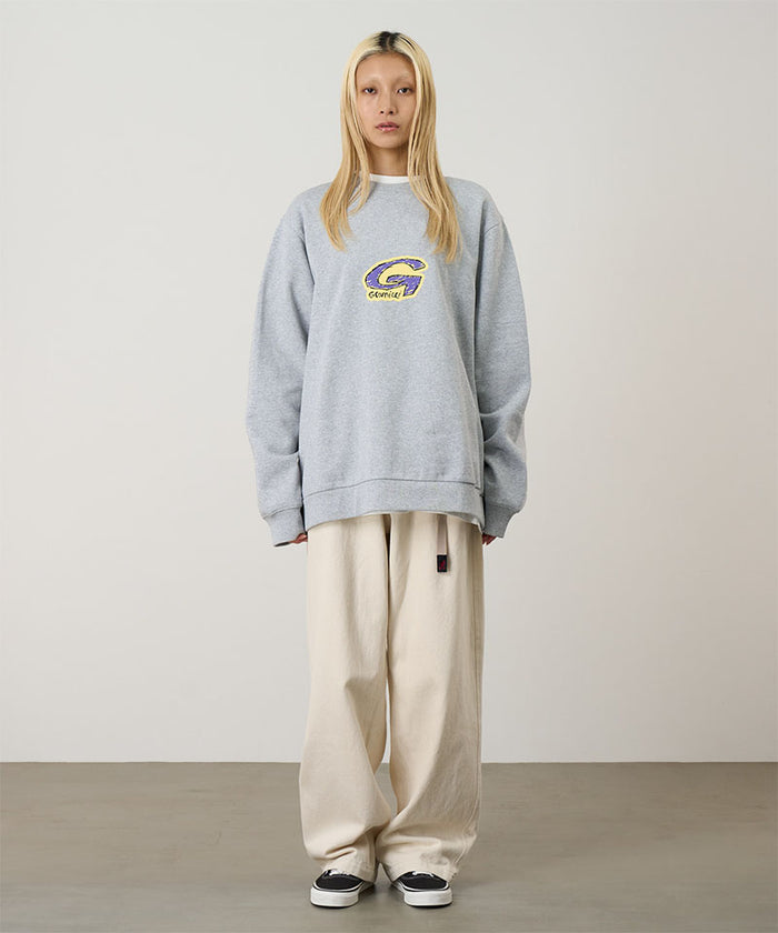 Model wearing a heather grey Gramicci G-Scribble Sweatshirt with a purple and yellow graphic and beige pants on a light tone background.