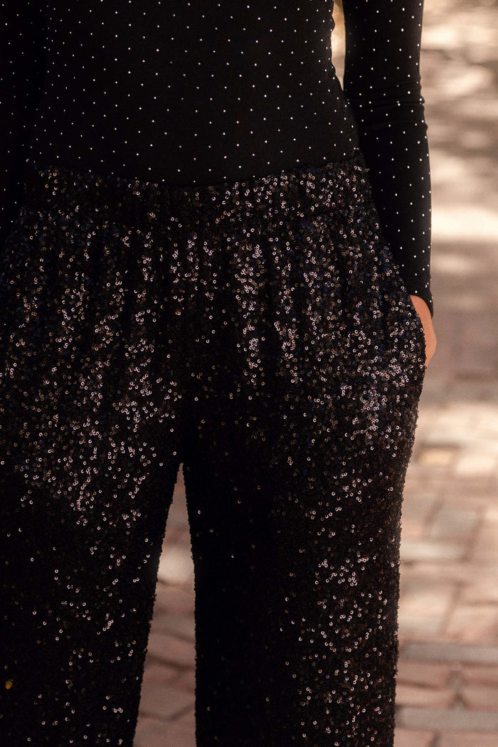 A person radiates Holiday Glam in a black, long-sleeve, polka-dot top and Ivy City Co Gloria Pants in Black Sequin, standing hands-in-pockets on a paved outdoor surface. This look is also Maternity Friendly.