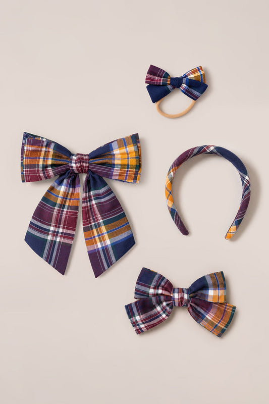 Four hair accessories from Ivy City Co’s Hair Bows in Midnight Plaid collection are displayed on a beige background: a large cotton bow, a medium Midnight Plaid bow on a French barrette clip, a small bow attached to an elastic band, and a matching headband—all in coordinating blue, yellow, orange, and burgundy tones.