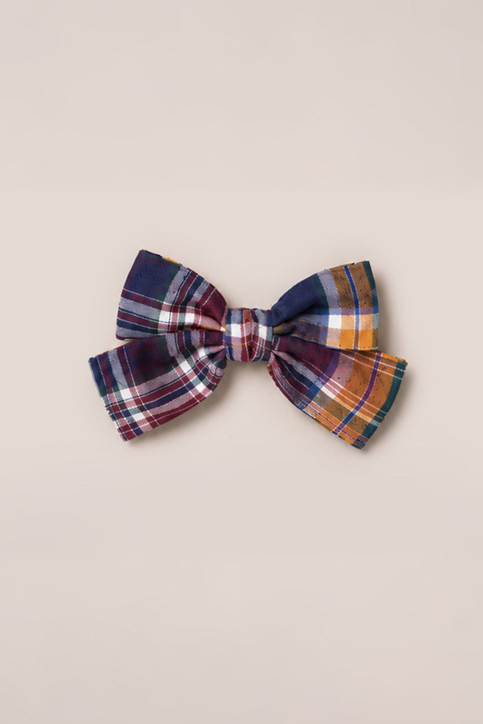 The Hair Bows in Midnight Plaid by Ivy City Co—a large accessory adorned with a classic plaid pattern in navy blue, burgundy, white, yellow, and orange—rests elegantly on a plain beige background, capturing timeless Ivy City charm.