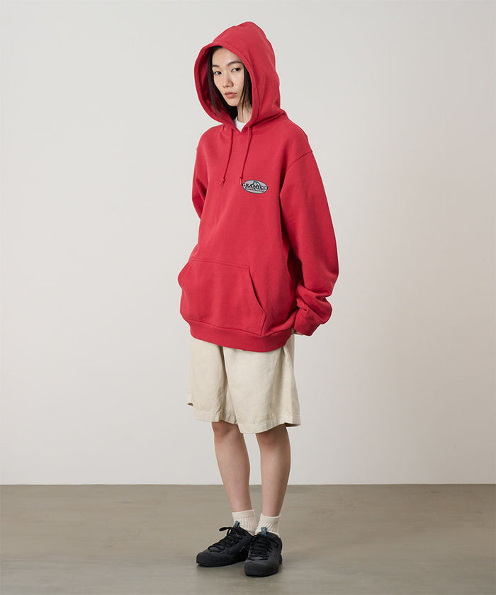 Model wearing a crimson red Gramicci Half Dome Hooded Sweatshirt with a beige and green graphic and beige shorts on a light tone background.