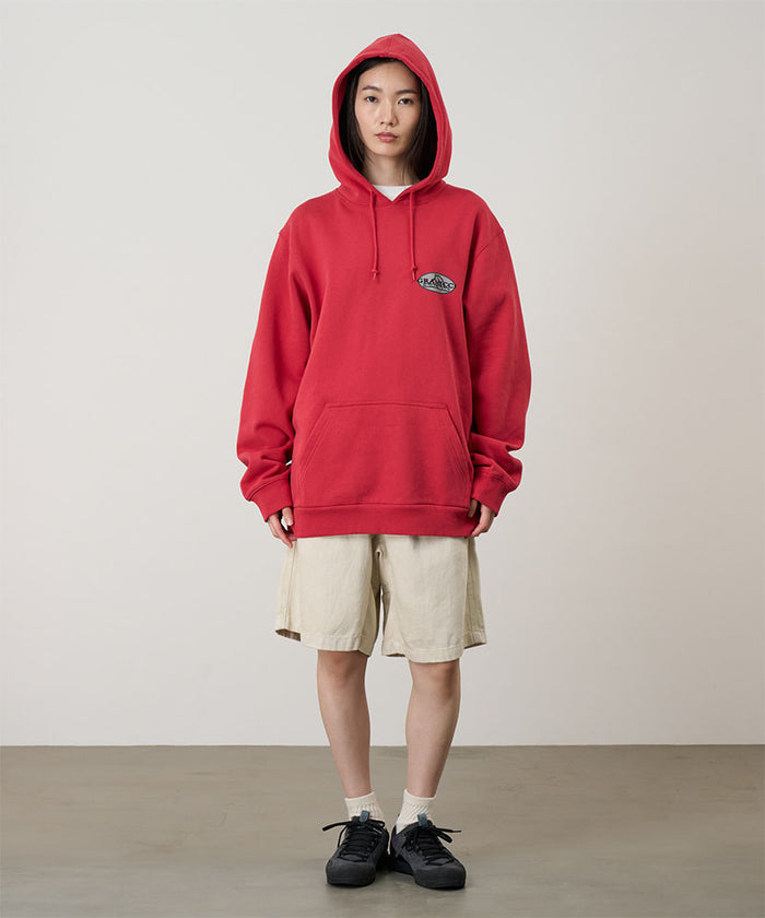 Model wearing a crimson red Gramicci Half Dome Hooded Sweatshirt with a beige and green graphic and beige shorts on a light tone background.