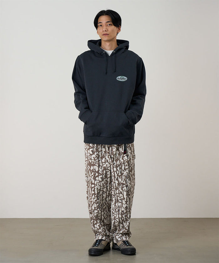Model wearing a vintage black Gramicci Half Dome Hooded Sweatshirt with a beige and green graphic and camouflage pants on a light tone background.