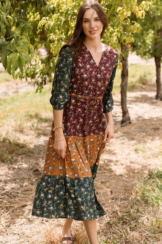 A woman stands outdoors among trees, wearing the Harlowe Dress in Elysian Vine by Ivy City Co—a long patchwork floral dress featuring maroon, green, and orange sections with elegant puff sleeves. Her long brown hair frames her face as she smiles softly, sunlight filtering through the leaves. The style is both charming and maternity friendly.