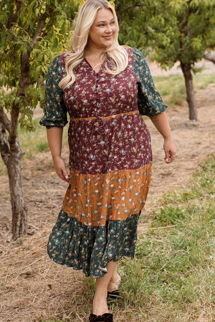 A woman with long blonde hair wears the Ivy City Co Harlowe Dress in Elysian Vine, a maternity-friendly patchwork floral dress with puff sleeves, as she walks outdoors along a tree-lined path, smiling and looking to the side.