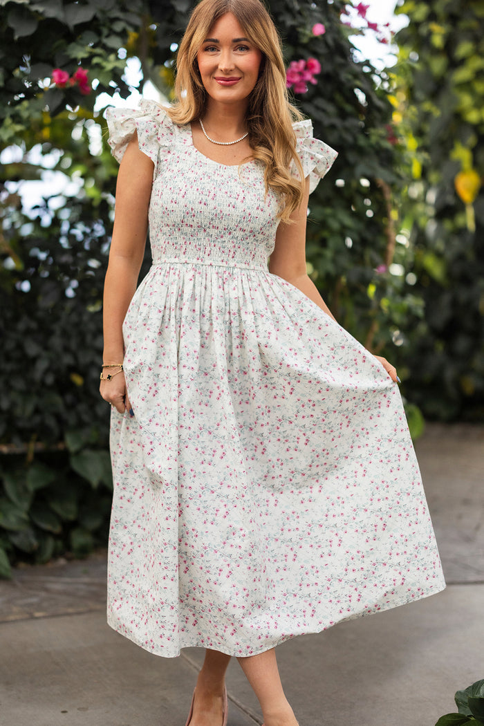 A woman outdoors smiles as she holds the sides of her Ivy City Co Hattie Dress in Pansy Trellis, a long white floral cotton dress with ruffled sleeves and a smocked bodice, perfect for nursing and maternity. She’s surrounded by lush plants and pink flowers.