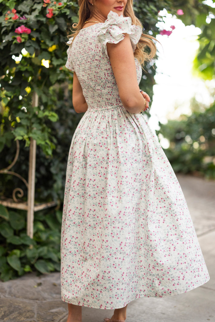A woman stands outdoors in the Ivy City Co Hattie Dress in Pansy Trellis, a light floral cotton dress with short ruffled sleeves. She’s turned slightly to the side among greenery and flowers. This style is nursing and maternity friendly.