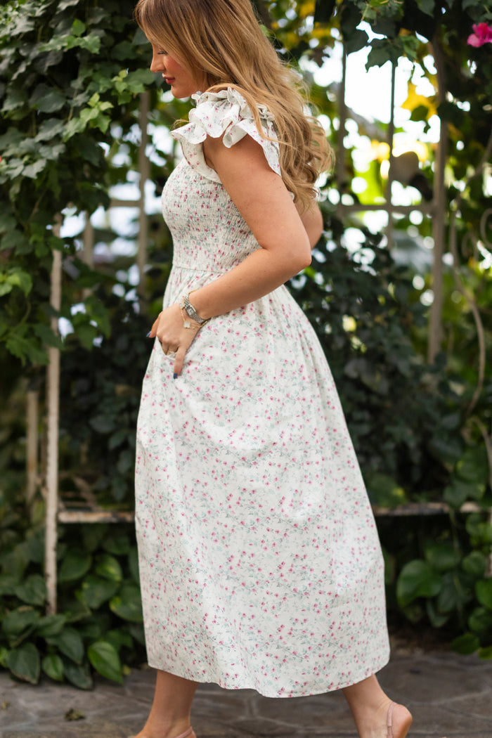 A woman with long, wavy hair walks outdoors in the Ivy City Co Hattie Dress in Pansy Trellis—a white, ankle-length floral cotton dress with ruffled sleeves that's nursing and maternity friendly—standing in profile amid green foliage.