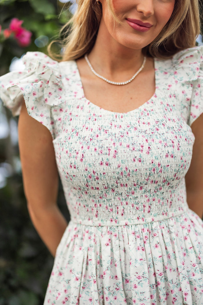 A woman in the Ivy City Co Hattie Dress in Pansy Trellis—a white floral cotton, nursing and maternity-friendly dress with pink prints and ruffled sleeves—stands outdoors, her wavy blonde hair and soft smile set against a blurred green background.