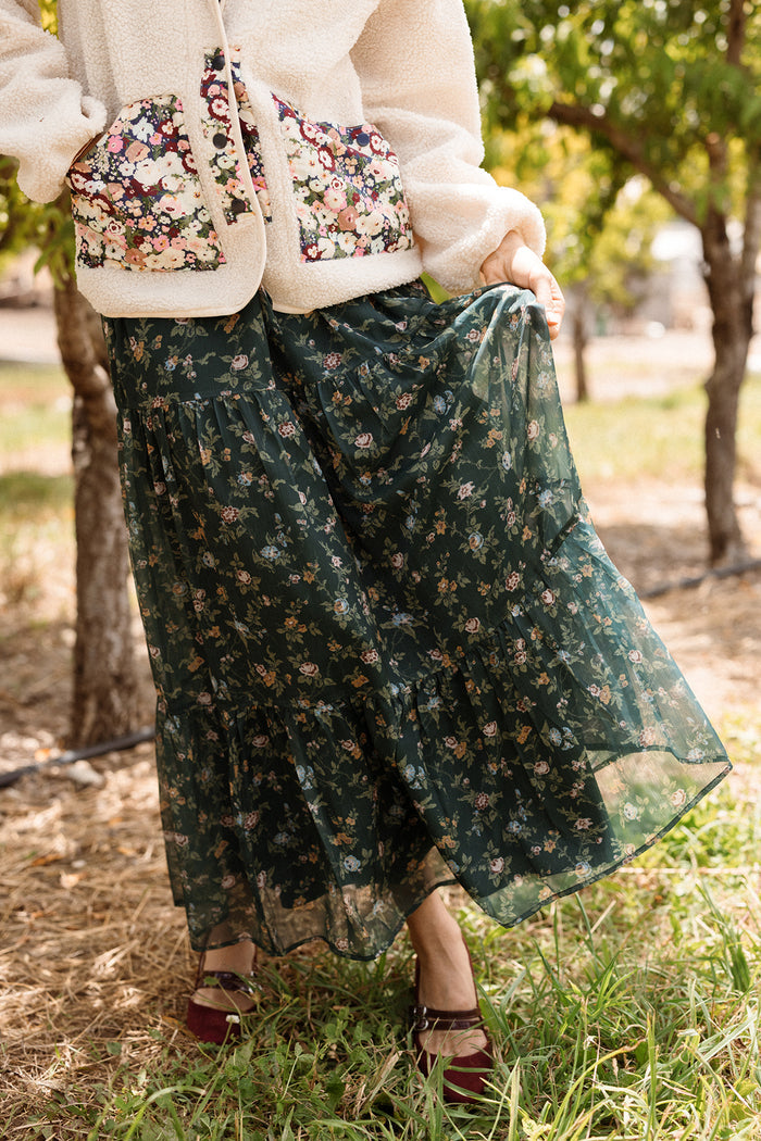 Outdoors among trees and grass, a person holds up the Ivy City Co Imogen Skirt in Evergreen Elysian Vine—a dark green skirt with a floral pattern—styled with a light jacket featuring floral pockets and burgundy shoes, for a perfect cozy fall look.