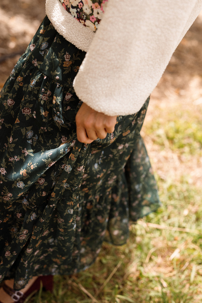 A person stands outdoors wearing a white sweater paired with the Imogen Skirt in Evergreen Elysian Vine by Ivy City Co, gently holding part of the skirt. Sunlight accentuates the elegant floral chiffon pattern and their hand, highlighting this beautiful piece as a charming addition to any fall wardrobe.