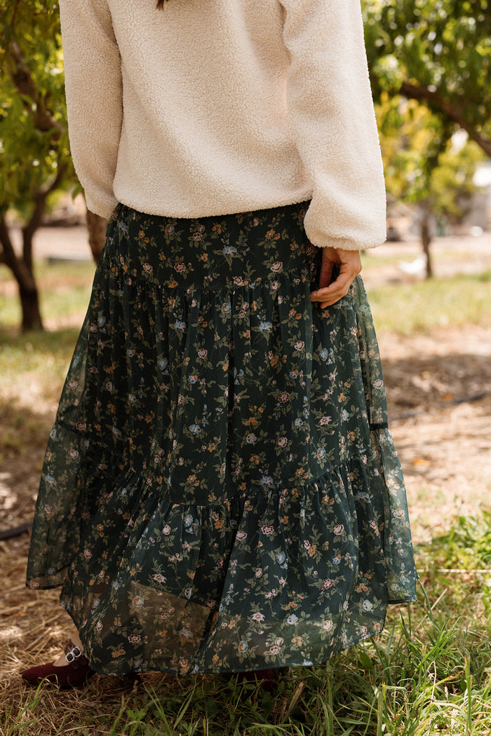 A woman wearing a cream sherpa sweatshirt and the Ivy City Co Imogen Skirt in Evergreen Elysian Vine, a dark green floral chiffon tiered maxi skirt, stands outdoors in a grassy area with trees in the background—an effortlessly cozy look perfect for your fall wardrobe.