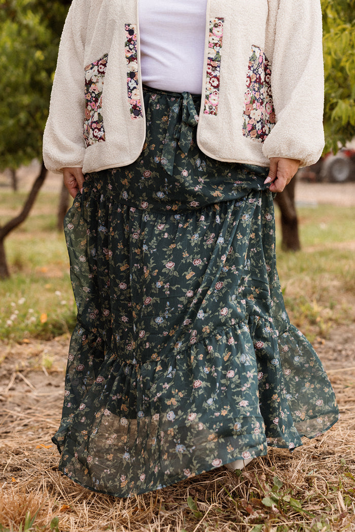 A person stands outdoors wearing a white jacket with floral pockets and the Ivy City Co Imogen Skirt in Evergreen Elysian Vine, featuring a dark green, floral-patterned design. With dry grass underfoot and trees in the background, it’s an ideal setting to showcase this stylish piece for your fall wardrobe.
