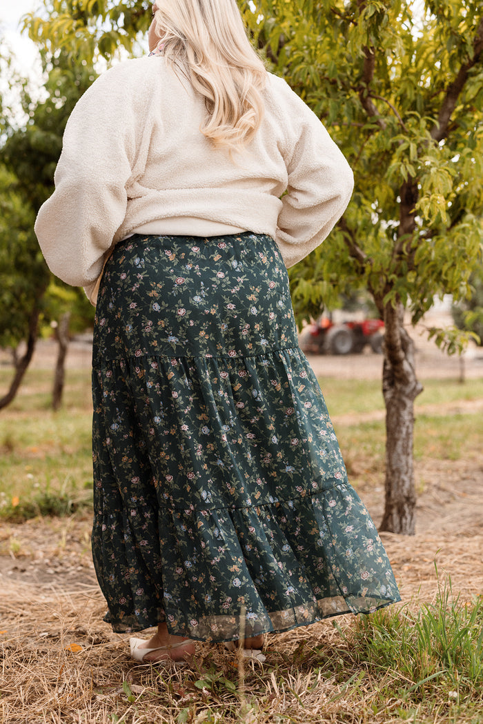 A person with long blonde hair, wearing a cream jacket and the Ivy City Co Imogen Skirt in Evergreen Elysian Vine, stands outdoors near trees with a red tractor in the background—capturing effortless style that’s perfect for any fall wardrobe.