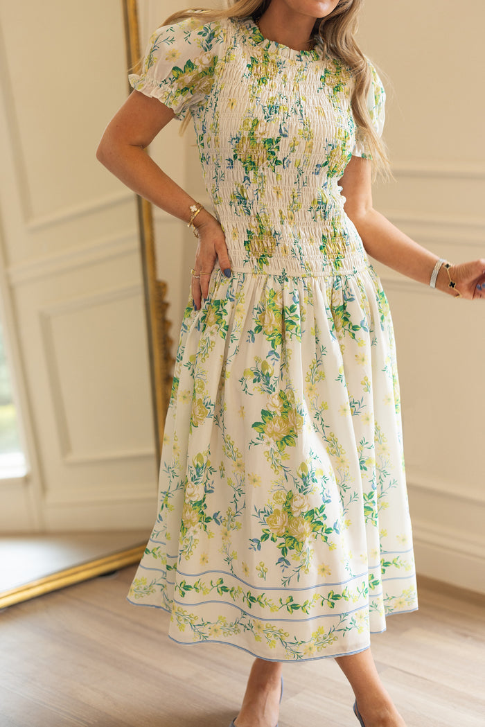 A woman wears the Ivy City Co Jennifer Dress in Garden Tea Party Floral, a white, short-sleeved, maternity-friendly dress with green and yellow blooms, standing indoors by a large gold-framed mirror.