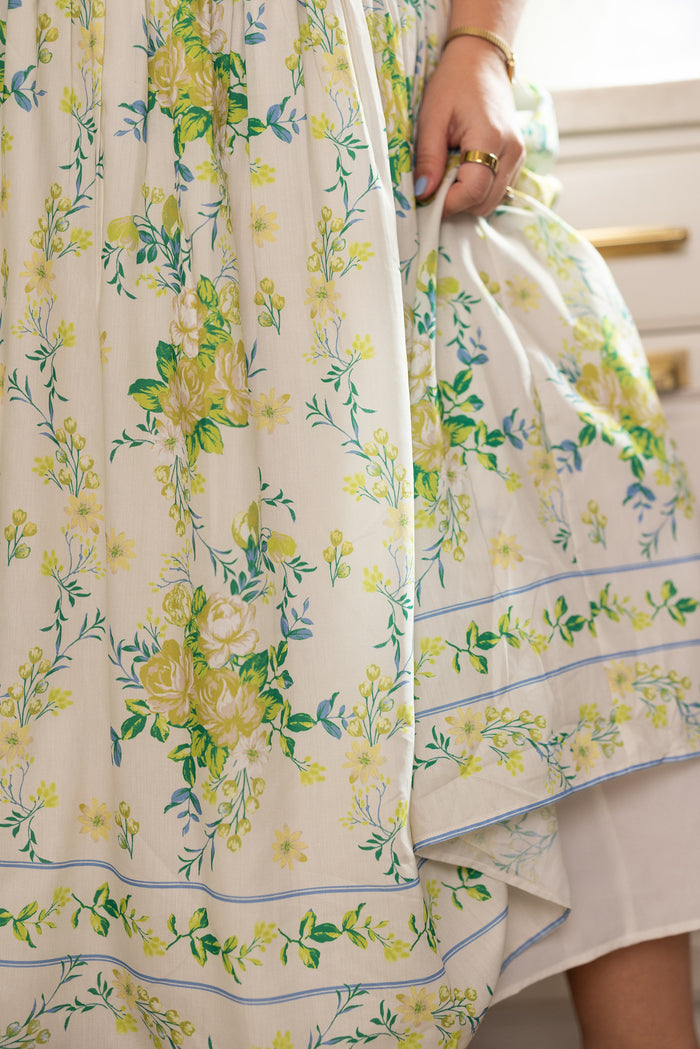 A close-up of Ivy City Co’s Jennifer Dress in Garden Tea Party Floral shows a hand with gold rings gently holding the white fabric, adorned with yellow and green floral details and blue accents. Drawer handles appear in the background.