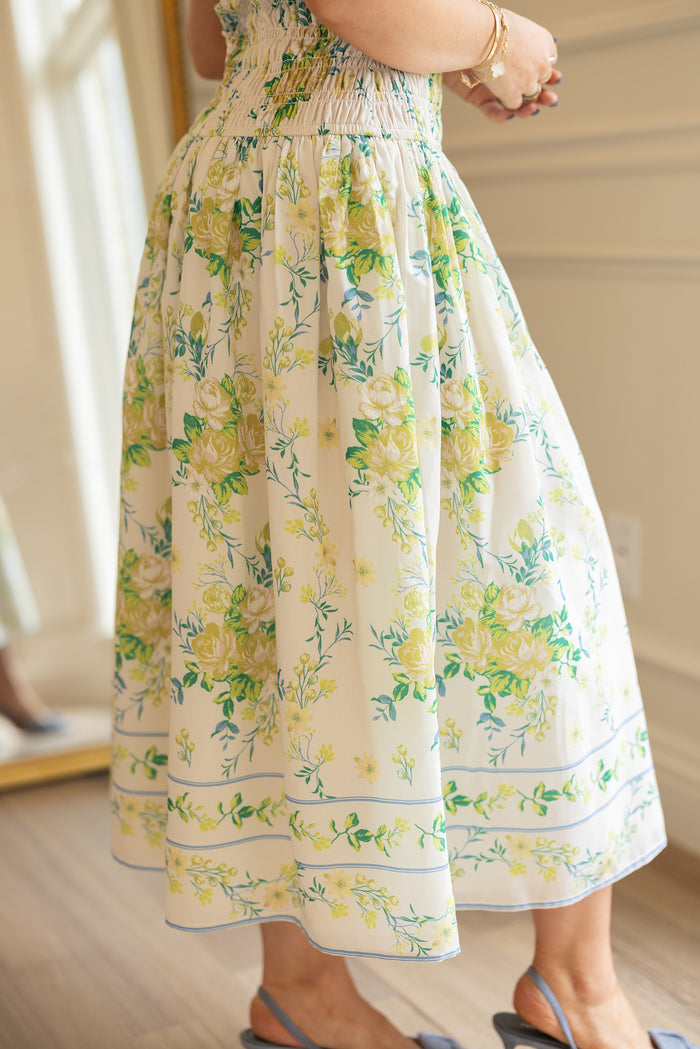 A person stands indoors on wooden flooring, wearing the Ivy City Co Jennifer Dress in Garden Tea Party Floral— a white, floral-patterned midi dress with yellow and green flowers—paired with blue sandals and hands clasped.