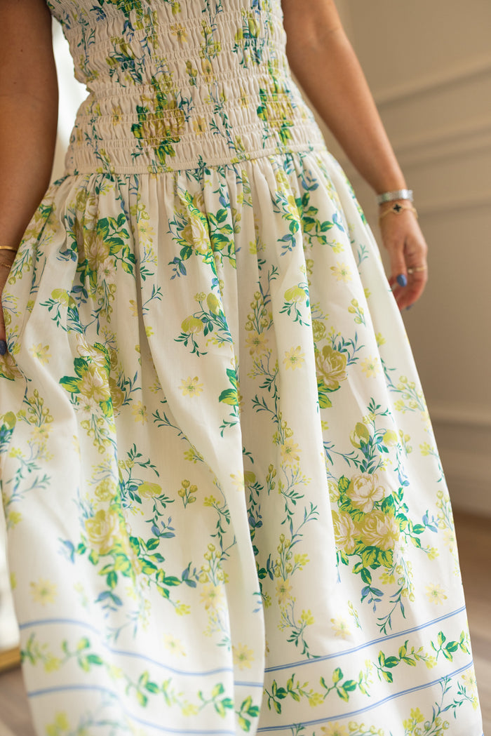 A person stands indoors wearing the Ivy City Co Jennifer Dress in Garden Tea Party Floral—a white, maternity-friendly dress with a smocked bodice and yellow, green, and blue florals.