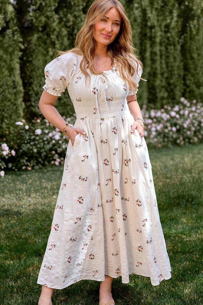 A woman stands on grass amid greenery and flowers, wearing the Ivy City Co Jessie Dress in Ditsy Embroidery—a white cotton dress featuring short puff sleeves and a small floral print.