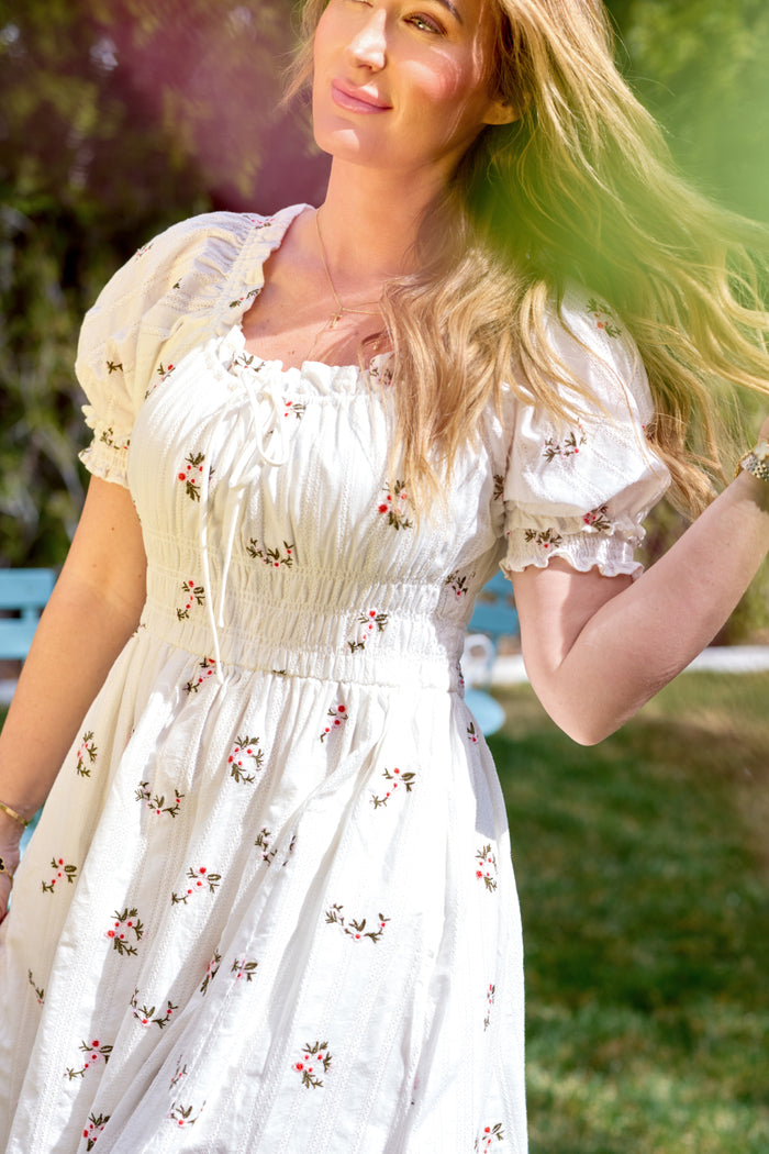 A woman wears the Ivy City Co Jessie Dress in Ditsy Embroidery, standing outdoors in sunlight with a hand raised. A green leaf is blurred in the foreground, with grass, trees, and a blue bench visible in the background.