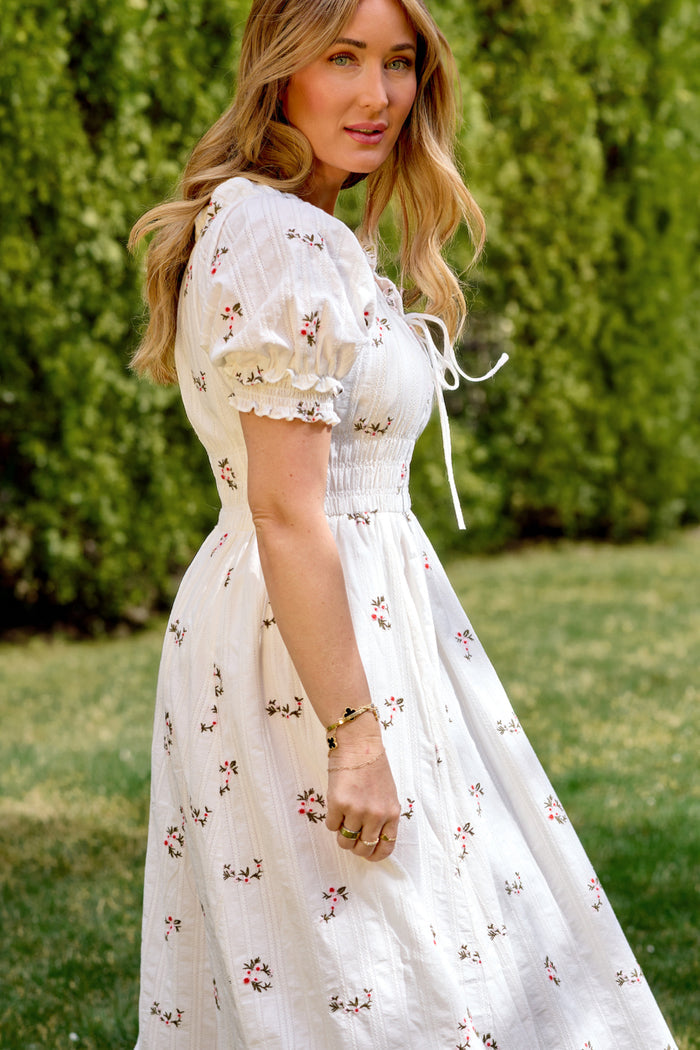 A woman with long, wavy blonde hair stands on grass in the Ivy City Co Jessie Dress in Ditsy Embroidery, a white cotton dress with small floral patterns and puffed sleeves. Green bushes are in the sunny background.