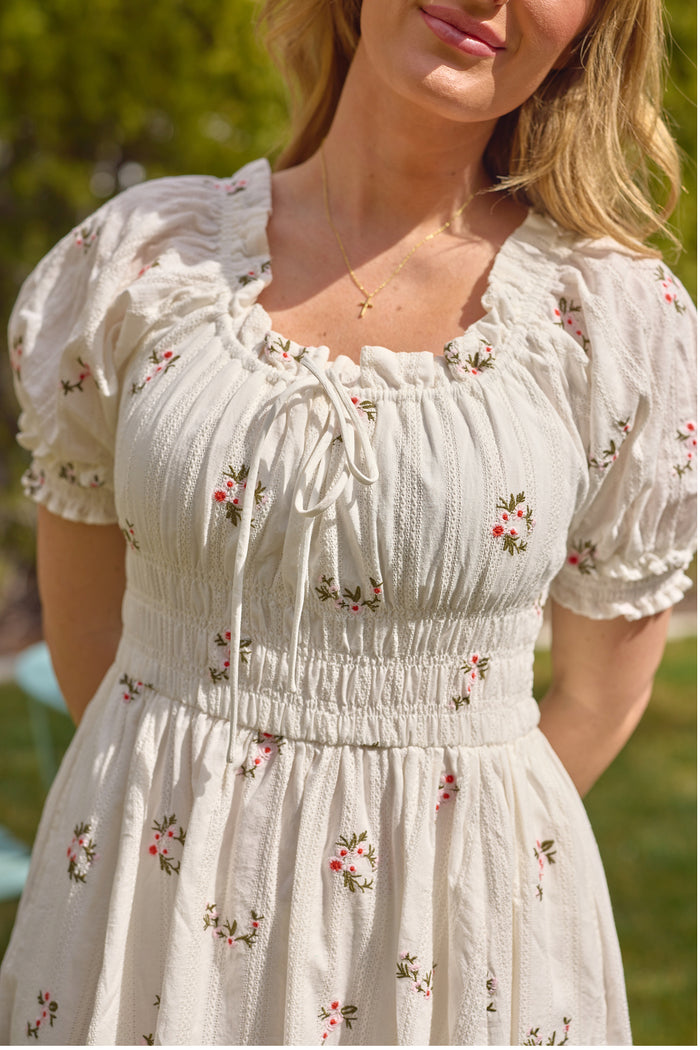 A woman with light hair wears the Ivy City Co Jessie Dress in Ditsy Embroidery—a light, short-sleeved, maternity-friendly dress with a ruched bodice and delicate floral detail—paired with a gold necklace outdoors against blurred greenery.