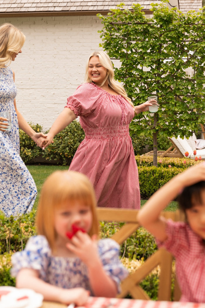Two women in Ivy City Co Jessie Dresses in Red Gingham hold hands and smile while walking through a garden. In the foreground, two children sit at a table eating strawberries, adding to the bright and cheerful scene.