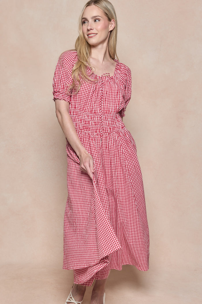 A woman wears the Ivy City Co Jessie Dress in Red Gingham, featuring puffed sleeves and a smocked bodice. She stands against a beige background, holding the skirt and smiling slightly. The style is maternity and nursing friendly.