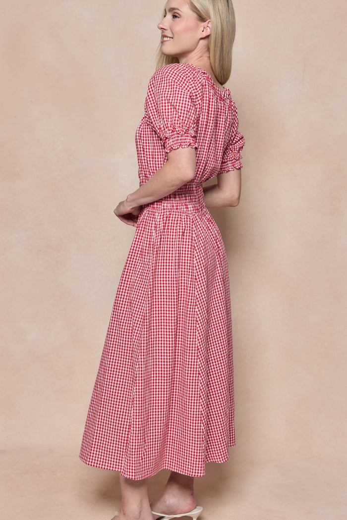 A woman with long blonde hair smiles sideways in the Ivy City Co Jessie Dress in Red Gingham, featuring a smocked bodice, fitted waist, short sleeves, and mid-calf length—maternity and nursing friendly—posed against a beige background.