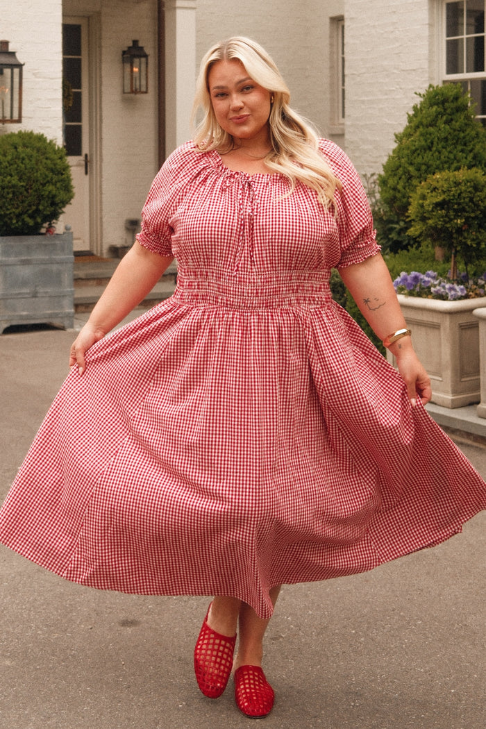 A woman with long blonde hair smiles outdoors, holding the sides of her Ivy City Co Jessie Dress in Red Gingham, with greenery and a white building in the background.
