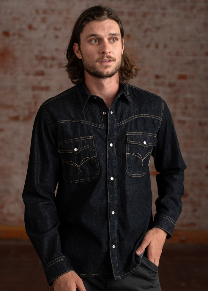 Man in a dark denim shirt with buttoned pockets stands against a brick wall, gazing to the side with a thoughtful expression.