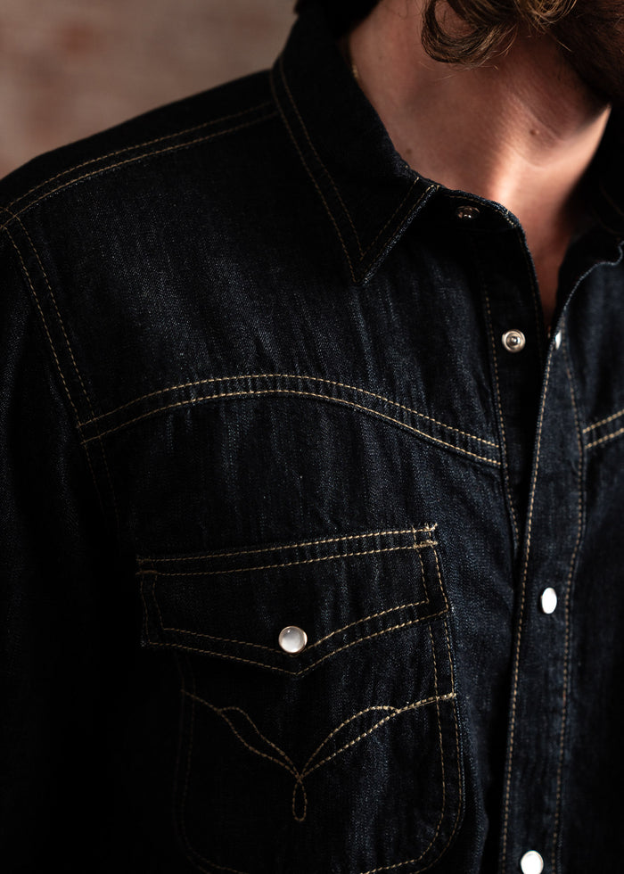 Close-up of a dark denim shirt with contrast stitching and snap buttons, highlighting the pocket and collar details.
