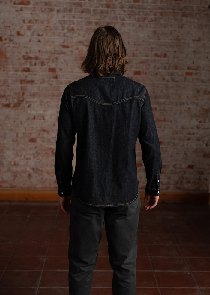 A person in a denim shirt and dark pants stands facing a brick wall, creating a rustic, casual atmosphere in the room.