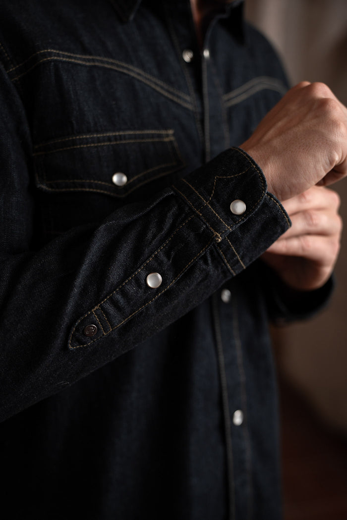A person adjusts the cuff of a dark denim shirt with white buttons, highlighting the detailed stitching and fabric texture.