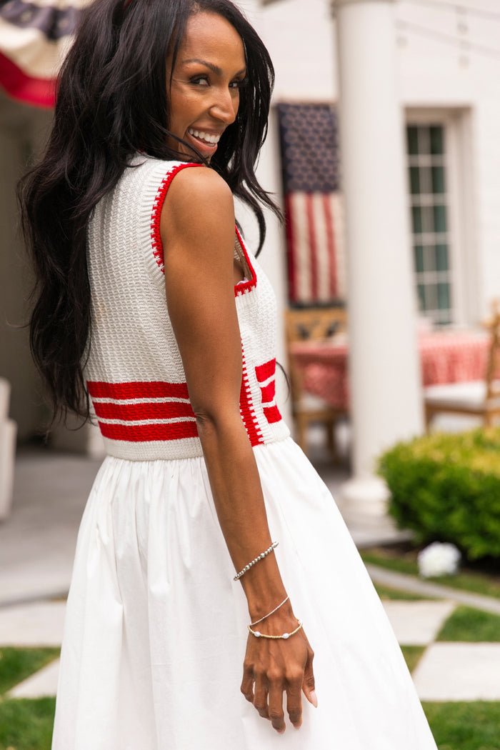 A woman with long dark hair smiles outdoors in the sleeveless Ivy City Co Lisbon Dress in Ivory & Firework Red. Crochet details and red stripes evoke a festive, patriotic vibe, perfect for the Fourth of July against an American flag backdrop.