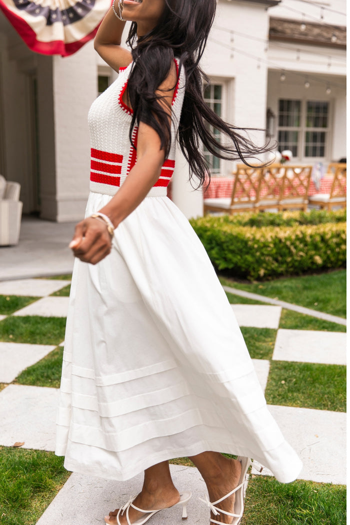 A woman twirls outdoors in the Ivy City Co Lisbon Dress in Ivory & Firework Red, her long dark hair flowing as she stands on a checkered stone path with garden furniture and greenery in the background.