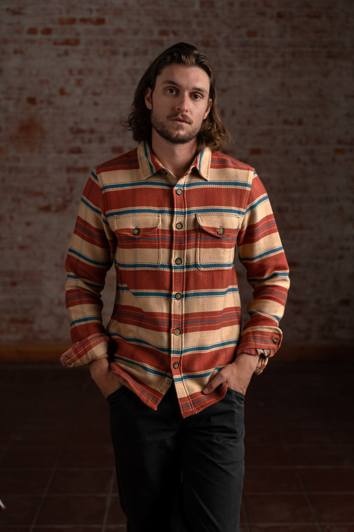 Man in a striped flannel shirt stands against a brick wall, hands in pockets, exuding a relaxed, casual vibe.